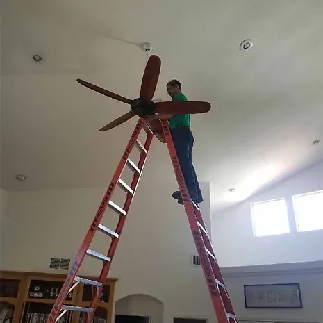 An electrician on a tall orange ladder installing a ceiling fan under a high ceiling